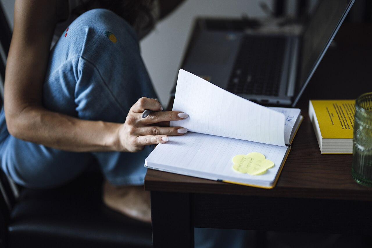Person writing notes in a notebook at a table with a laptop and books nearby